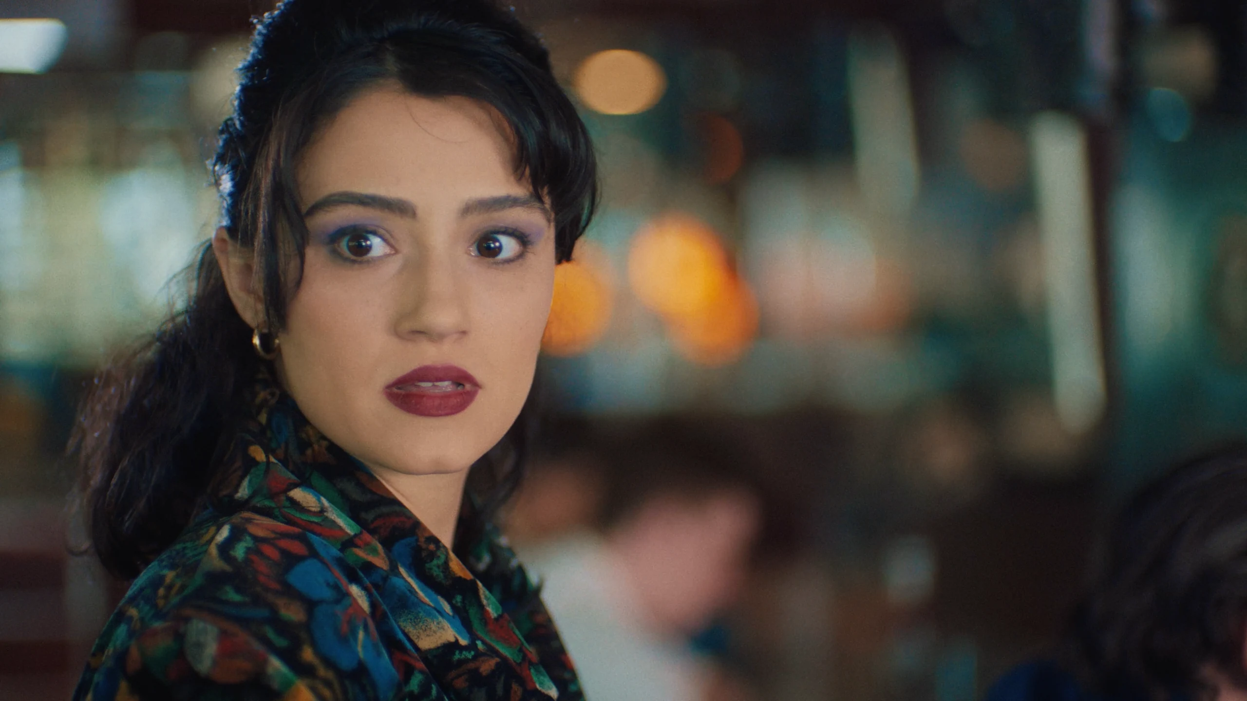 Close-up of a wide-eyed woman with dark hair, burgundy lipstick and a patterned jacket against a blurred, warmly lit interior