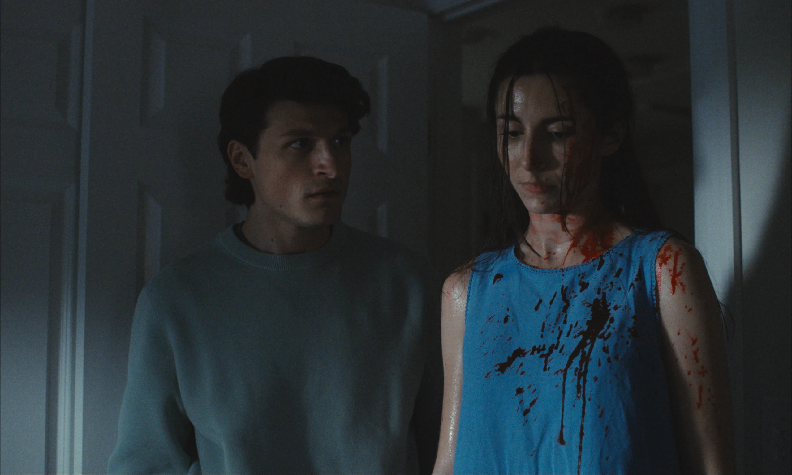 In dim light, a man in a pale sweater looks toward a wet-haired woman with blood on her face and blue top