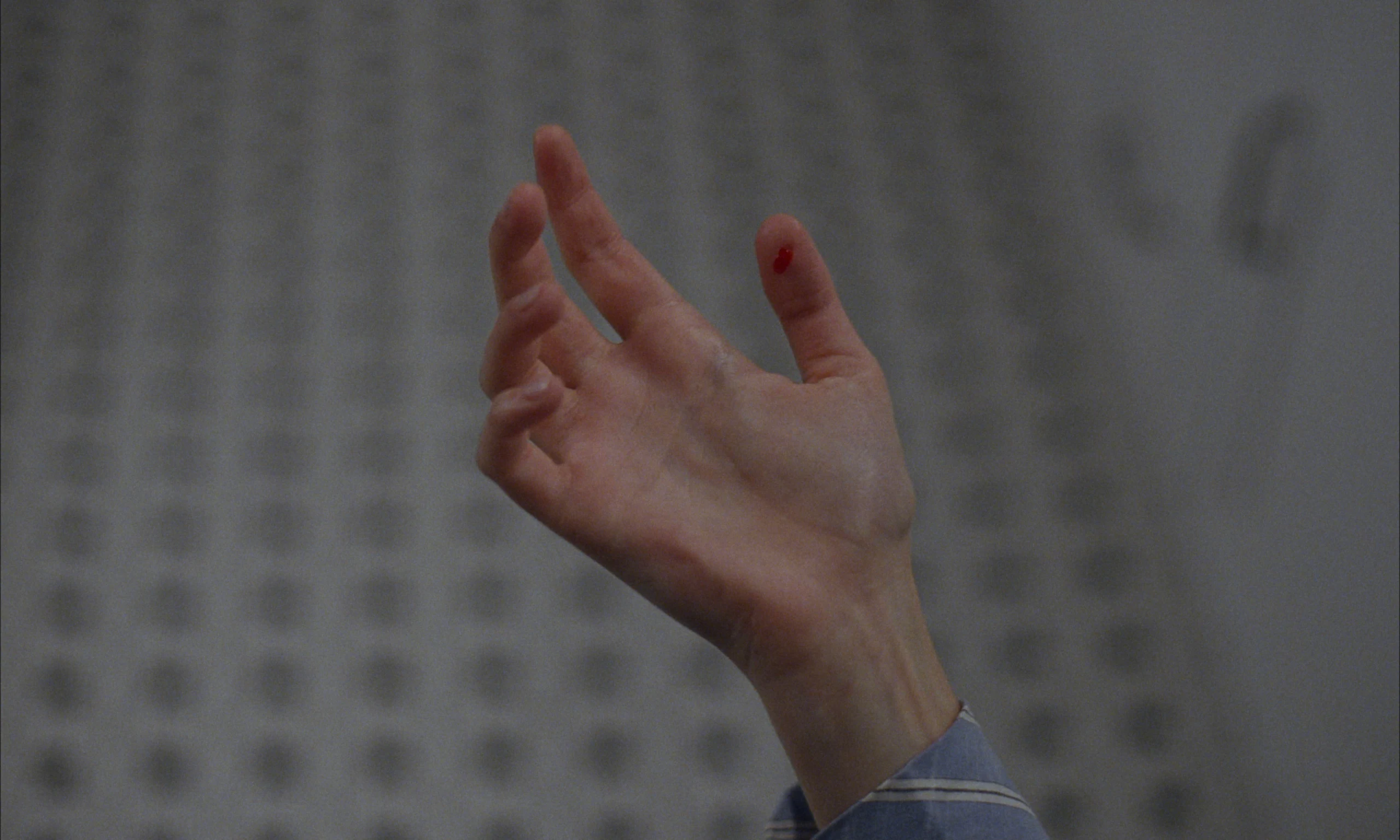 A raised open hand with a small red spot on the thumb, against a blurred grey patterned wall