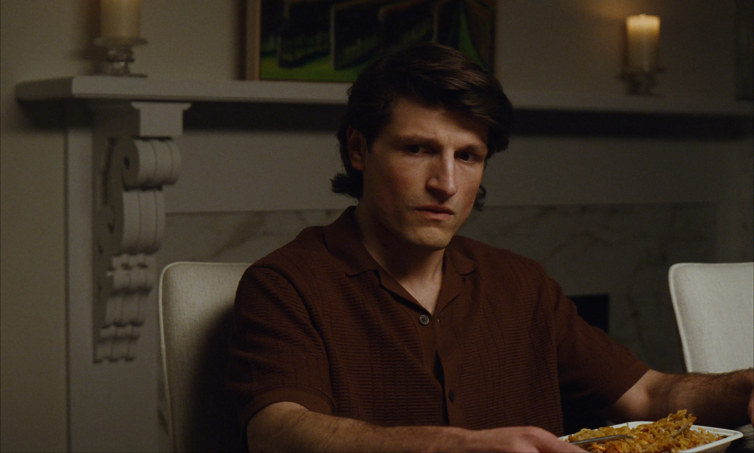 A man with collar-length dark hair in a brown shirt sits at a table with noodles, candles and a mantel behind