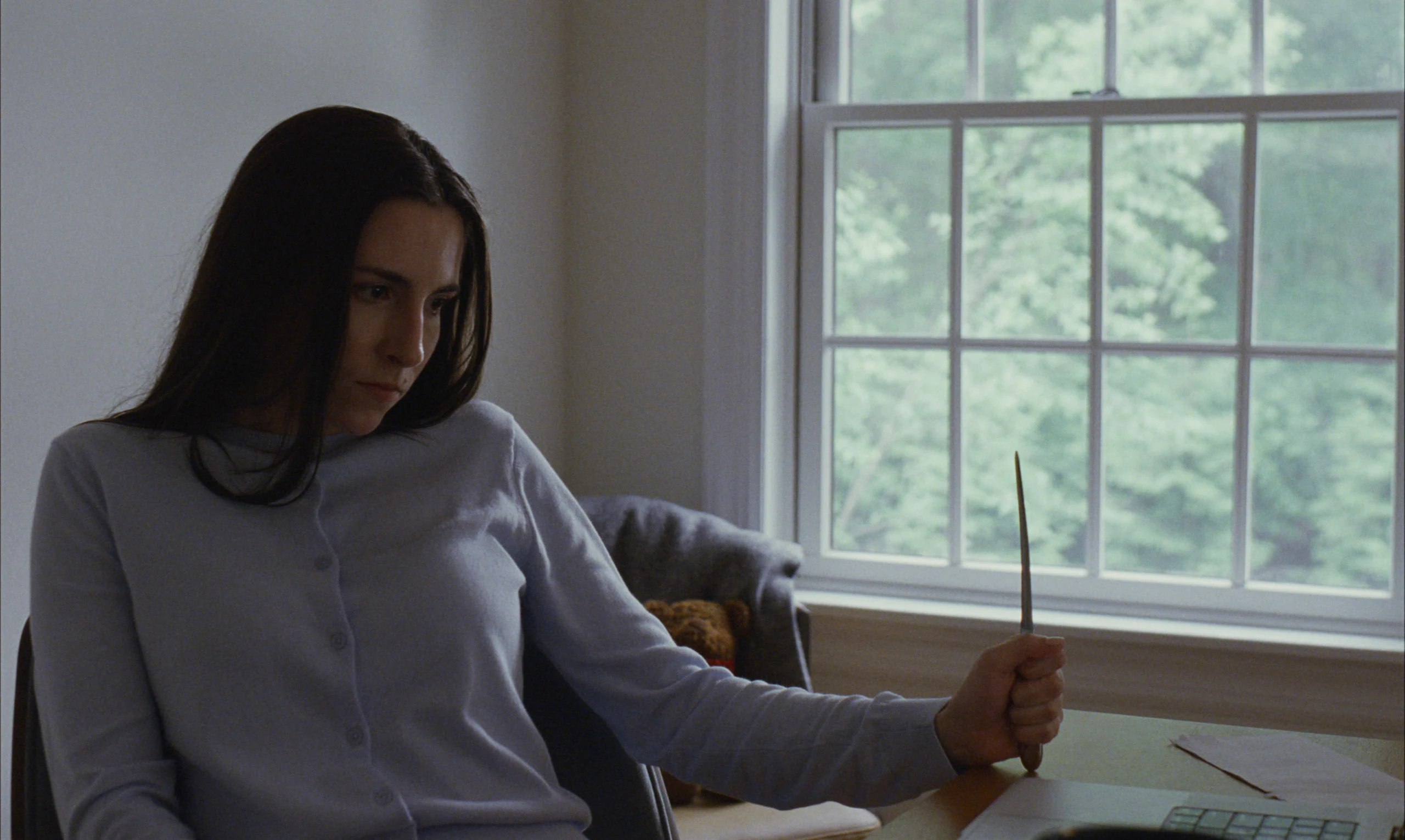 A woman with long dark hair in a pale blue cardigan sits by a window, holding a knife upright on a desk