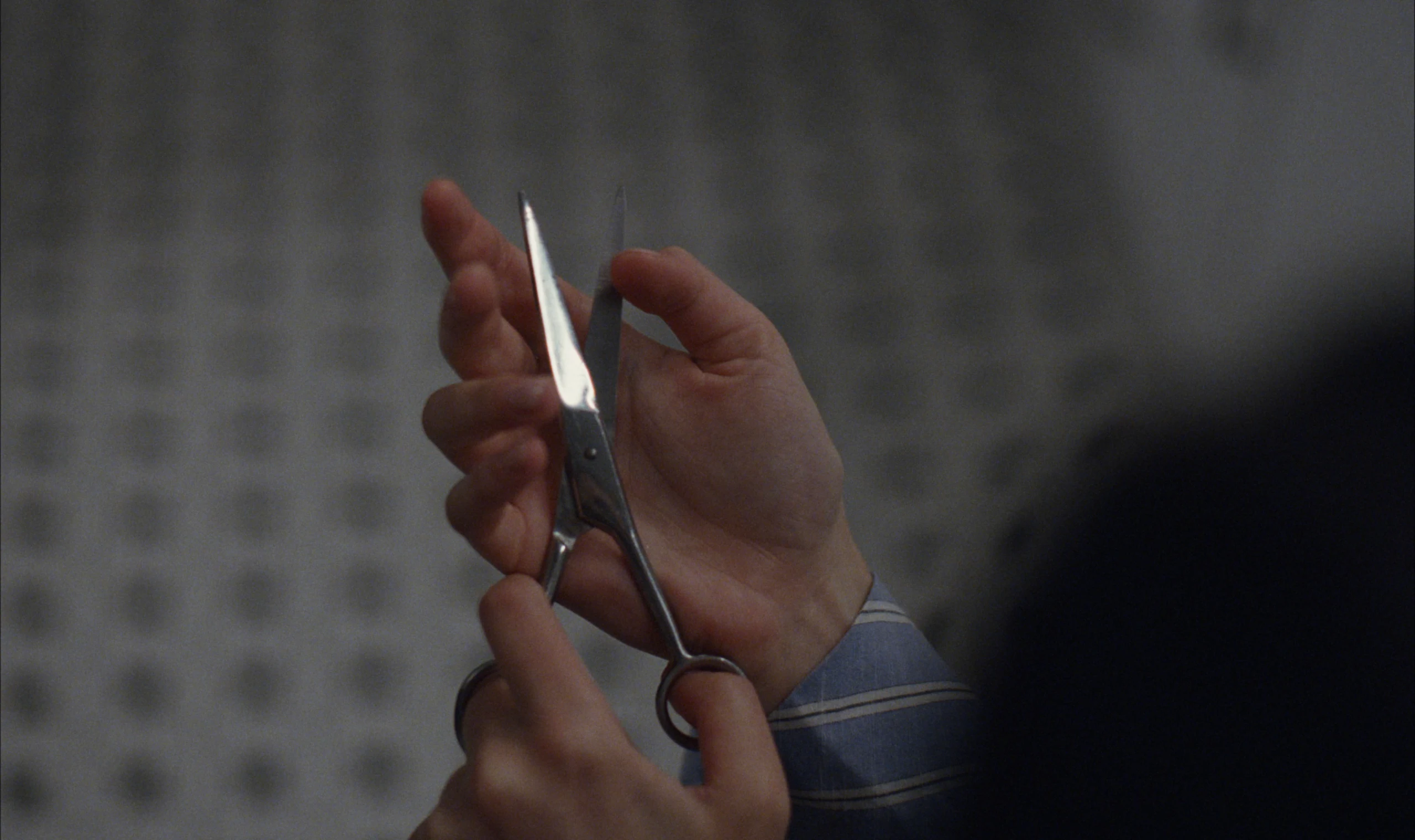 Two hands, one at a striped blue shirt cuff, open a pair of silver scissors against a blurred patterned wall