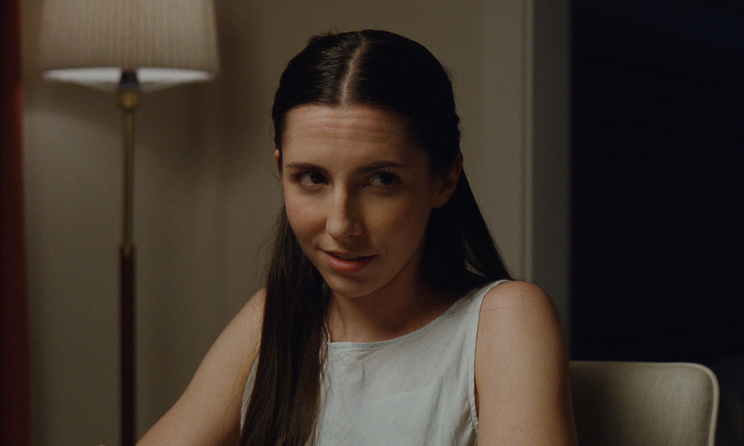 A woman with long dark hair and a white sleeveless top smiles and glances sideways, a lit floor lamp behind her