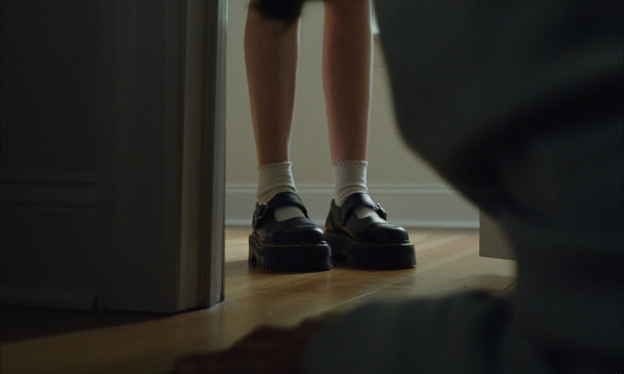 Low view through a doorway of legs in white frilled socks and black platform Mary Jane shoes on a wooden floor