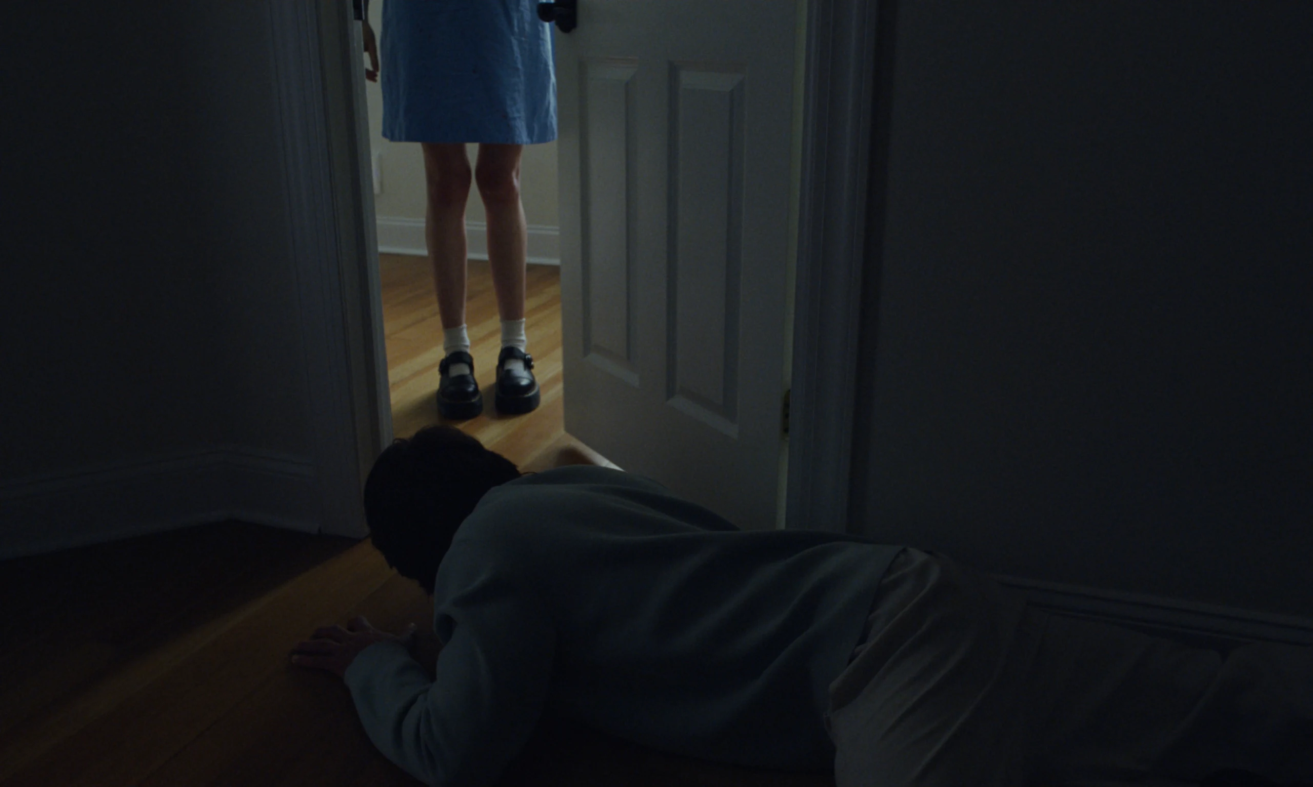 A short-haired figure in a pale sweater lies prone on the floor as someone in a blue dress stands in the doorway