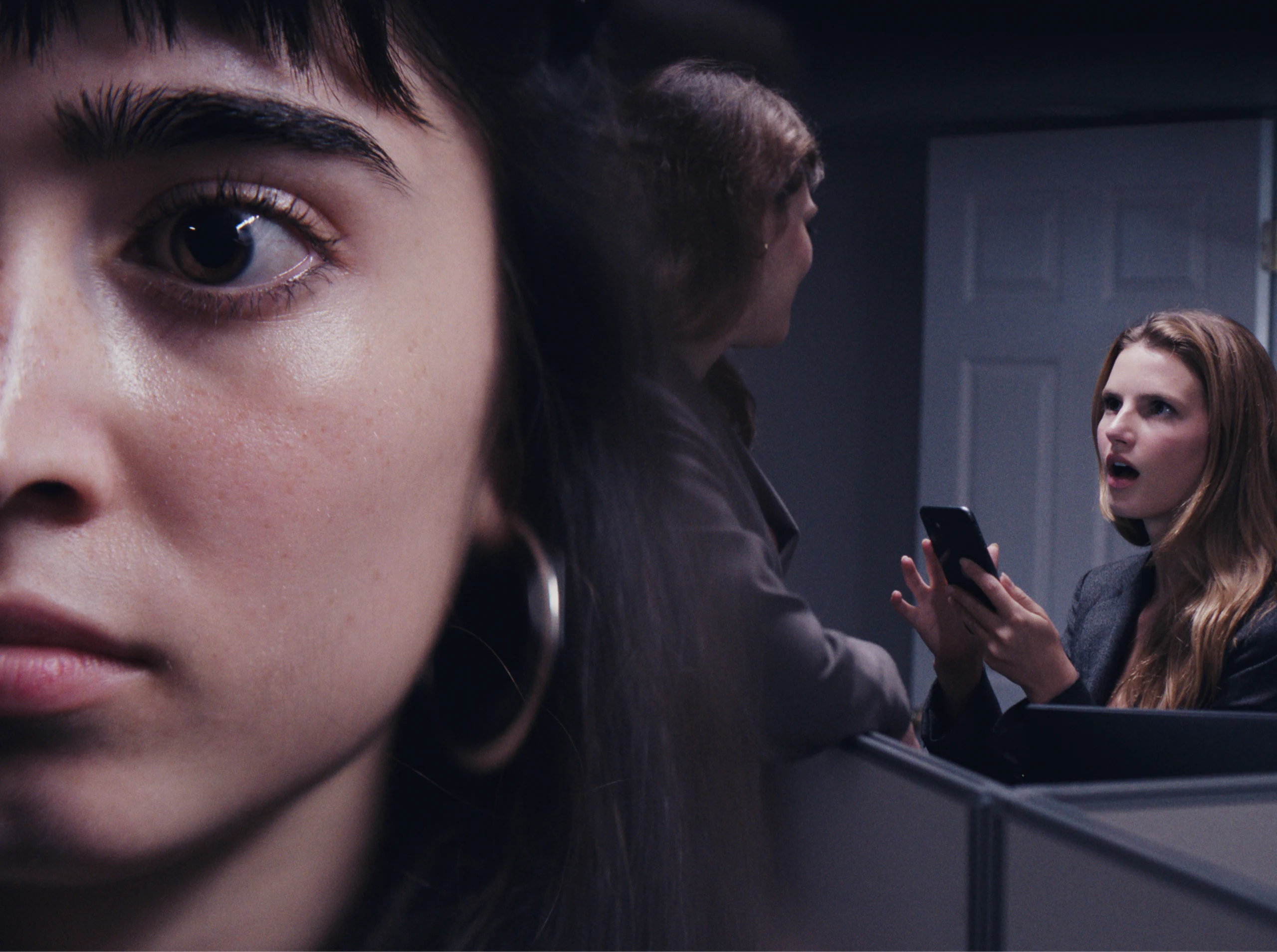 A dark-haired woman with bangs fills the left of frame while a woman with a phone stares open-mouthed across an office cubicle