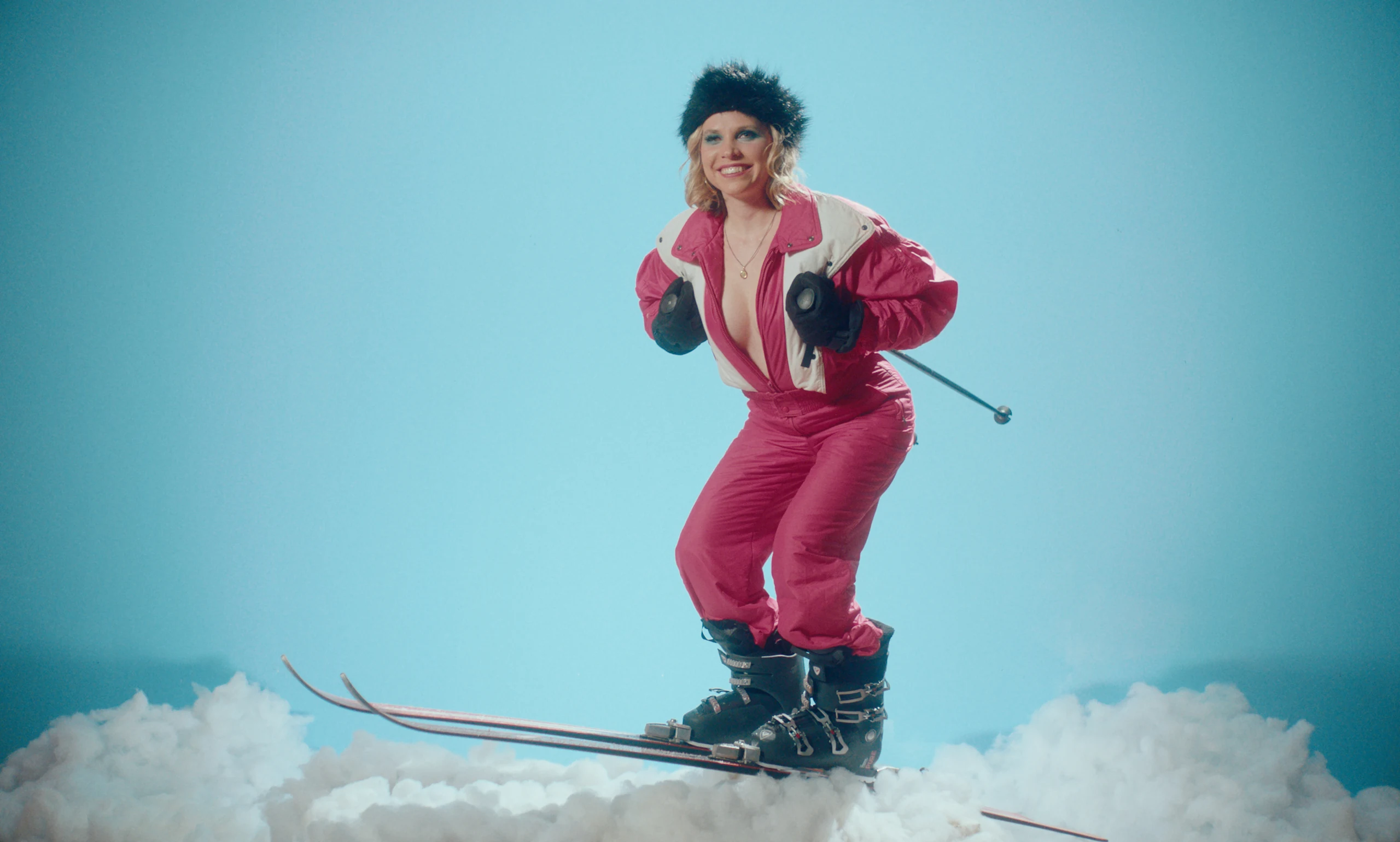 A smiling woman in a pink ski suit and black fur hat crouches on skis over cotton snow against pale blue