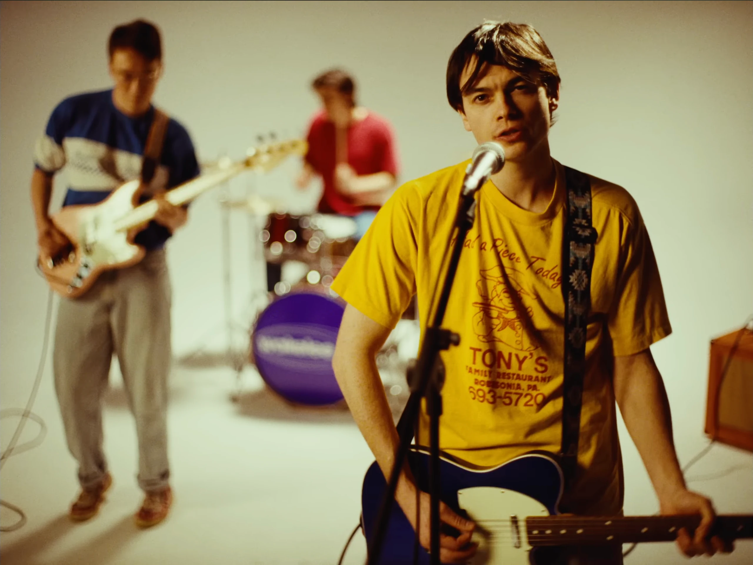 A singer in a yellow shirt plays guitar at a microphone, with a bassist and drummer behind him in a white studio.